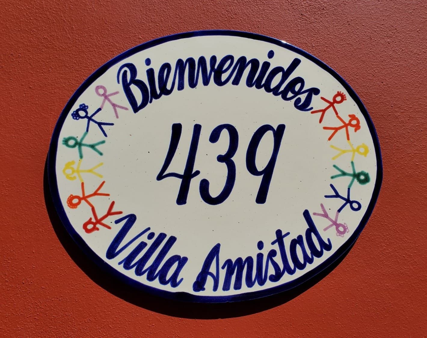 El letrero de cerámica pintado a mano en el muro de terracota de la villa: Bienvenidos, 439, Villa Amistad, rodeado de figuritas tomadas de la mano