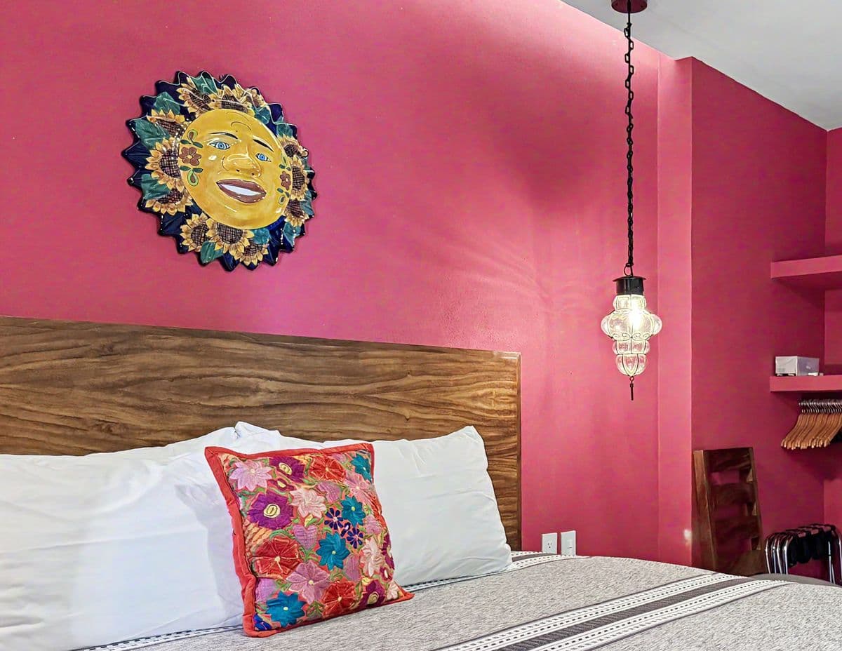 Una cama king en Ivan's Bella Vista: almohadas blancas, un cojín mexicano bordado y un sol de Talavera en la pared rosa de arriba